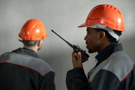 Young adult man using walkie talkie communicating with colleague while working on construction site, both wearing safety helmets and uniforms, standing against plain backgroundの写真素材