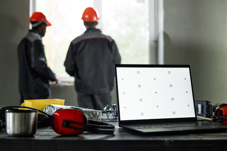 Two middle aged men wearing safety helmets standing near window discussing construction project, foreground showing open laptop with blank screen and protective headphonesの写真素材
