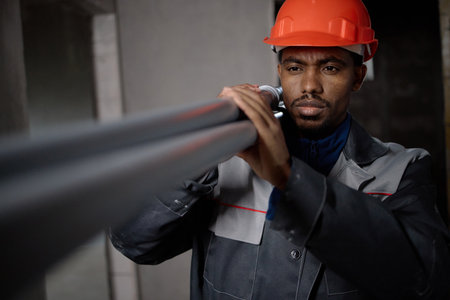 Black young adult man wearing hard hat installing metal pipes in industrial setting, focusing intently on alignment, demonstrating skilled manual labor in construction environmentの写真素材