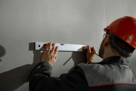 Middle aged Caucasian man wearing hard hat using spirit level and pencil marking measurements on wall, focusing on precise alignment during construction or renovation workの写真素材