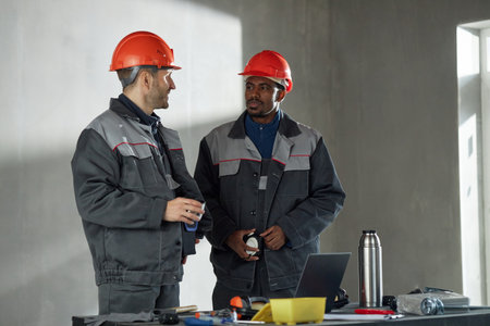 Two middle aged men, one Caucasian and one Black, wearing protective helmets and work uniforms, standing at table with tools and laptop, discussing project in industrial settingの写真素材