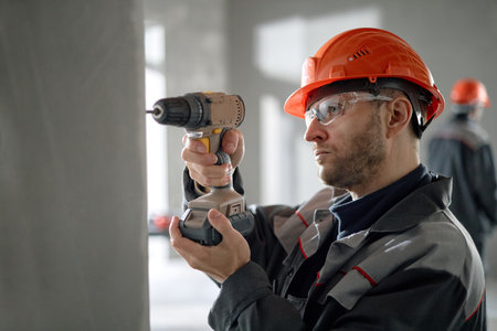 Caucasian middle aged man wearing safety helmet and protective glasses operating cordless power...