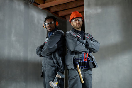 Two young adult men, one Black and one Caucasian, standing back to back with arms crossed, wearing protective workwear and safety gear, posing confidently at construction siteの写真素材