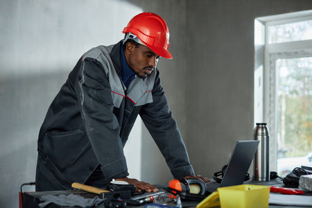 Black man wearing protective helmet and work uniform standing at table using laptop, appearing focused while reviewing construction plans or technical documents in industrial settingの写真素材