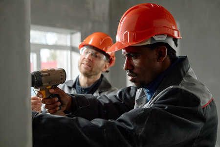 Black man using electric drill while middle aged Caucasian man supervising in unfinished building, both wearing protective helmets and work jackets, focused on construction taskの写真素材