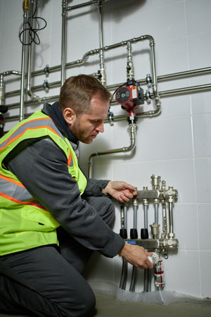 Middle aged Caucasian man kneeling while adjusting industrial plumbing valves on wall, wearing...