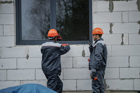Two middle aged men wearing protective helmets working together outside unfinished building, one man pointing at window frame while other holding construction toolの写真素材