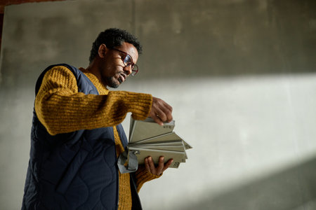 Young adult man wearing glasses holding and examining color swatches in hands standing against plain industrial background looking focusedの写真素材