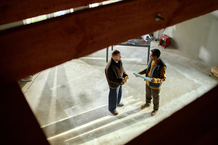 Caucasian middle aged man and Black middle aged man standing in unfinished room discussing construction project, holding blueprint, sunlight streaming through windows onto concrete floorの写真素材