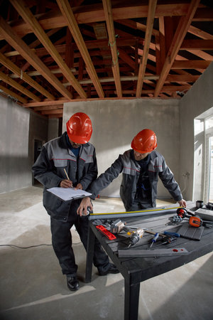 Two middle aged men wearing safety helmets reviewing construction plans and measuring materials on table inside unfinished building with exposed wooden beamsの写真素材
