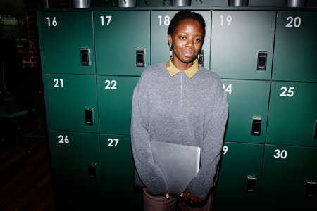 Portrait of young adult Black woman standing in front of green lockers holding closed laptop, smiling slightly, wearing casual office attire in modern business office settingの写真素材