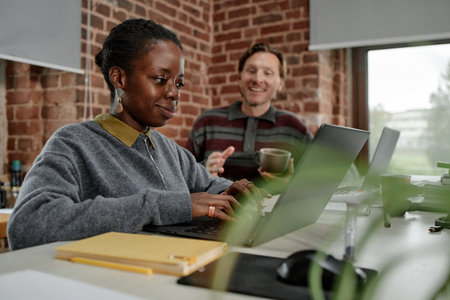 Young adult Black woman working on laptop while middle aged Caucasian man standing nearby, holding coffee cup and smiling in modern business office with exposed brick wallsの写真素材