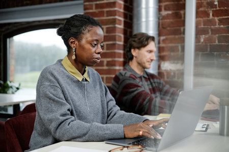 Black young adult woman working on laptop beside Caucasian middle aged man typing on computer in...