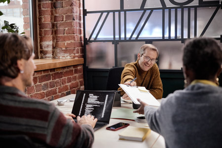 Young adult Caucasian man smiling while handing document to middle aged Black woman during business meeting, young adult Caucasian man using laptop with code on screen in modern officeの写真素材