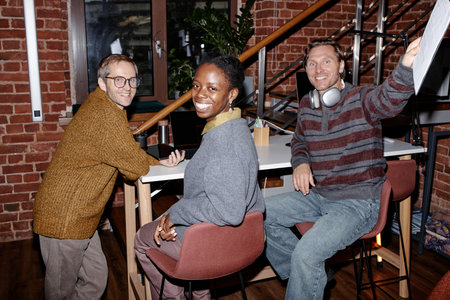 Three smiling colleagues, young Caucasian man, Black woman, middle aged Caucasian man, collaborating in modern business office, sitting and standing near high table, casual interactionの写真素材