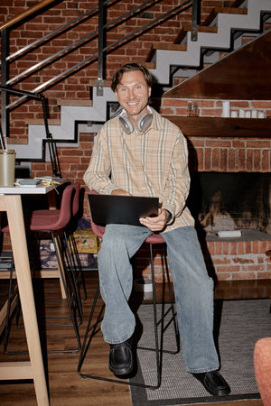 Portrait of middle aged Caucasian man sitting on high chair holding laptop and wearing headphones...