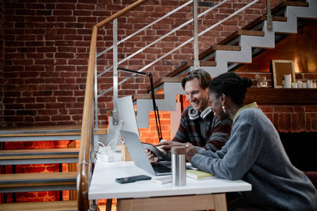 Caucasian middle aged man and Black young adult woman collaborating at computer workstation in modern business office, both focused on screen and discussing project togetherの写真素材