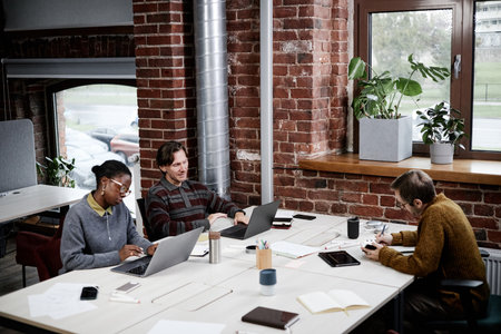 Diverse group of young adult and middle aged colleagues working on laptops and writing notes in modern business office, Black woman typing, Caucasian men focused on tasks, brick wall in backgroundの写真素材