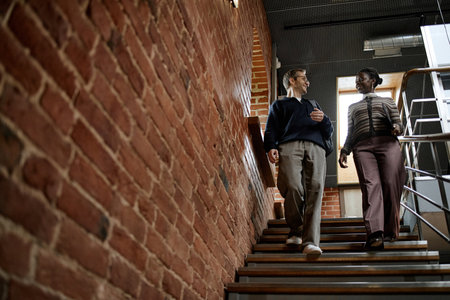 Middle aged Caucasian man and middle aged Black woman walking down staircase in modern business...
