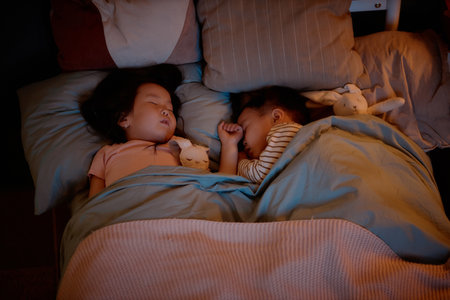 Asian children sleeping side by side in bed, young girl and toddler boy resting under blanket with stuffed animals, siblings sharing peaceful moment during evening at homeの写真素材