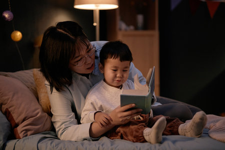 Asian middle aged woman sitting on bed reading book to Asian child in evening home setting, both...