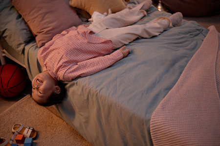 Asian child lying upside down on bed smiling, spending evening at home, visible basketball and toys...