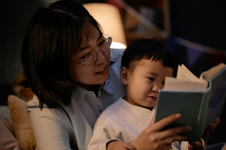 Asian middle aged woman sitting with Asian child reading book together in living room during evening, both focusing on pages, woman supporting child while sharing storyの写真素材