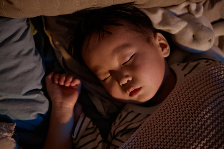 Asian child sleeping peacefully on bed with head resting on pillow, eyes closed, hand near face, surrounded by soft bedding and plush toy, wearing striped shirt, nighttime settingの写真素材