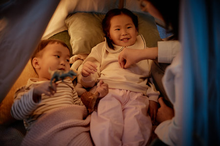 Asian woman interacting with two Asian children lying together under blanket, young boy holding toy dinosaur, all smiling and engaging in playful activity during evening at homeの写真素材