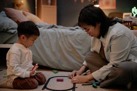 Asian young woman sitting on floor assembling toy train track with child holding toy, spending...