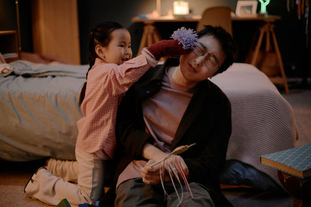 Asian child playfully touching face of Asian young adult man with fuzzy toy while kneeling on floor in bedroom, man holding handmade craft, both engaging in playful family interactionの写真素材