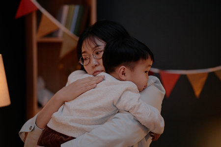 Asian middle aged woman hugging Asian child indoors during evening, both facing sideways, woman wearing glasses, child resting head on womans shoulder, festive bunting in backgroundの写真素材