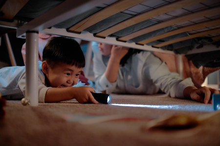 Asian child boy lying on floor under bed holding flashlight, smiling and playing with mother, family spending evening together at home, engaging in playful activityの写真素材