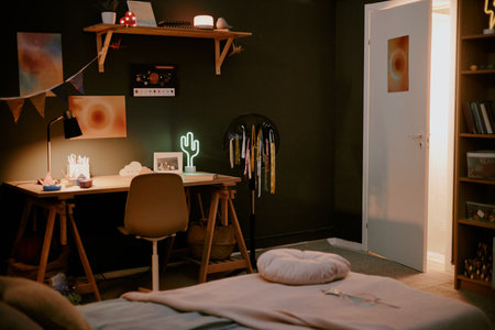 Cozy bedroom scene showing empty workspace desk with chair, illuminated lamp, open door revealing light from hallway, bed in foregroundの写真素材