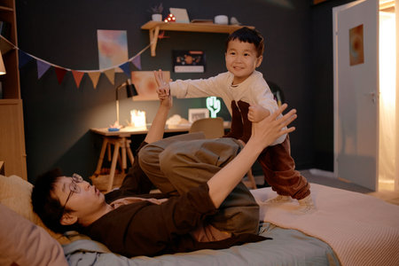 Asian middle aged man lying on bed holding hands with Asian child standing and smiling, spending...