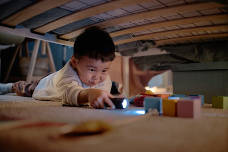 Asian child lying on floor under bed reaching with flashlight toward colorful toy blocks, smiling...