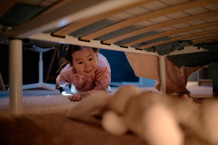 Asian child girl crawling on carpet under bed reaching toward plush toy smiling, evening indoor home setting, playful momentの写真素材