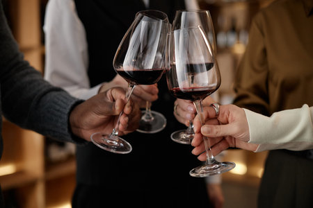 Diverse group of young adult and middle aged men and women clinking wine glasses during wine tasting, hands visible holding glasses with red wine, multiethnic participants engaging in social activityの写真素材