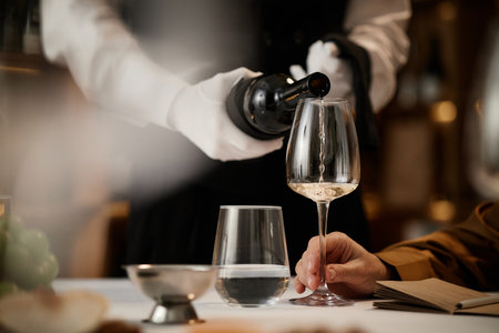 Caucasian man holding wine glass while sommelier in uniform pouring white wine during wine tasting session, closeup of hands and glassware on table, focus on tasting processの写真素材