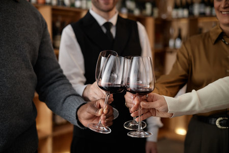 Diverse group of young adult and middle aged men and women clinking wine glasses during wine tasting, hands holding glasses with red wine, shelves with bottles in backgroundの写真素材