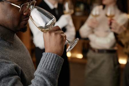 Black man smelling wine in glass during tasting session with two Caucasian young adults holding wine glasses in background, group participating in wine evaluation eventの写真素材