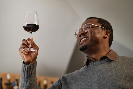 Black middle aged man holding wine glass up to light, examining color and clarity while smiling, participating in wine tasting process in indoor setting, wearing eyeglassesの写真素材