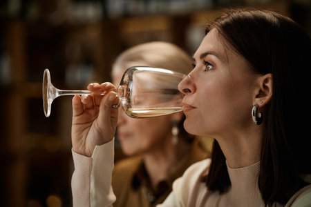 Young adult Caucasian woman tasting wine in profile with middle aged Caucasian woman in background participating in wine testing session, both focusing on evaluating flavor and aromaの写真素材