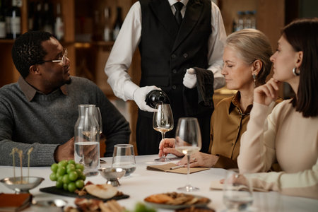 Middle aged Black man, senior Caucasian woman, young adult Caucasian woman sitting at table watching waiter pouring wine into glass, people participating in wine tasting event indoorsの写真素材