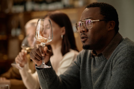 Middle aged Black man holding wine glass and examining color while sitting beside Caucasian woman and senior Caucasian woman, group participating in wine tasting session indoorsの写真素材