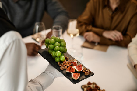 Group of middle aged multiethnic adults sitting at table tasting wine, holding glasses while server presenting slate platter with green grapes, figs and assorted nuts in foregroundの写真素材