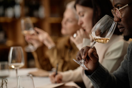 Diverse group of young adult and middle aged people tasting wine, holding glasses and evaluating aroma during wine testing session, focus on Black man smelling wine in foregroundの写真素材
