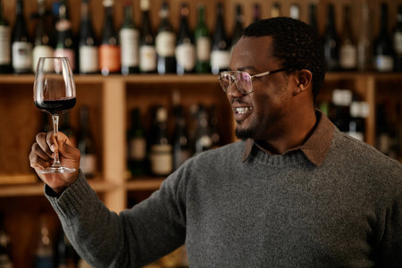 Black middle aged man holding wine glass, examining color and clarity of red wine in front of wine shelves, smiling while testing wine quality in professional tasting settingの写真素材