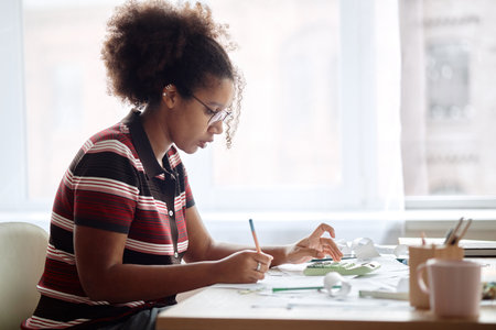 Young Black woman analyzing invoices and bills at desk, holding pencil and reviewing financial documents, focused on paperwork with calculator and office supplies nearbyの写真素材