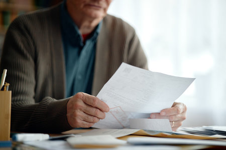 Senior man analyzing invoices and bills at desk, holding documents in hands, focusing on financial paperwork, managing personal expenses in home office environmentの写真素材